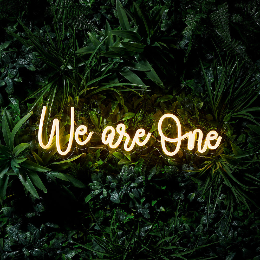 Leuchtender LED Neon Schriftzug We are One vor dunklem Pflanzenhintergrund
