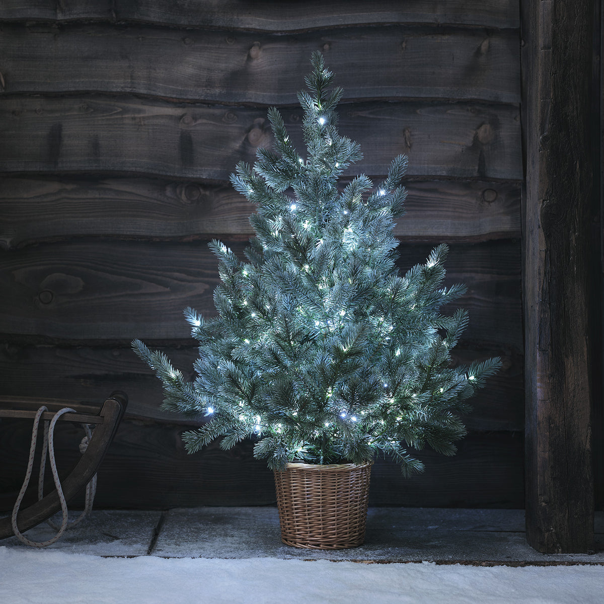 Weihnachtsbaum mit kaltweißen LEDs im Rattan-Korb neben Holzstuhl und Geschenken vor hellgrauer Wand