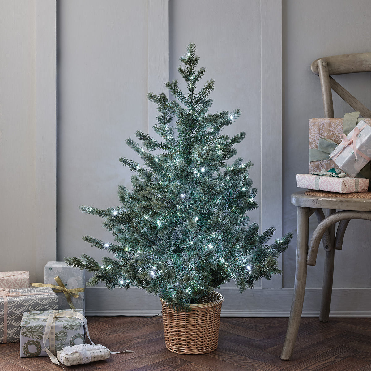 Weihnachtsbaum mit kaltweißen LEDs im Rattan-Korb vor hellgrauer Wand neben Holzstuhl und Geschenken