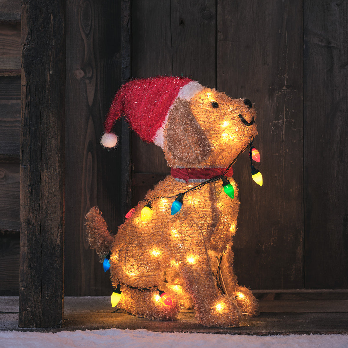 LED Weihnachtsfigur Welpe mit roter Weihnachtsmütze und bunter Lichterkette im Maul vor dunklem Holzhintergrund