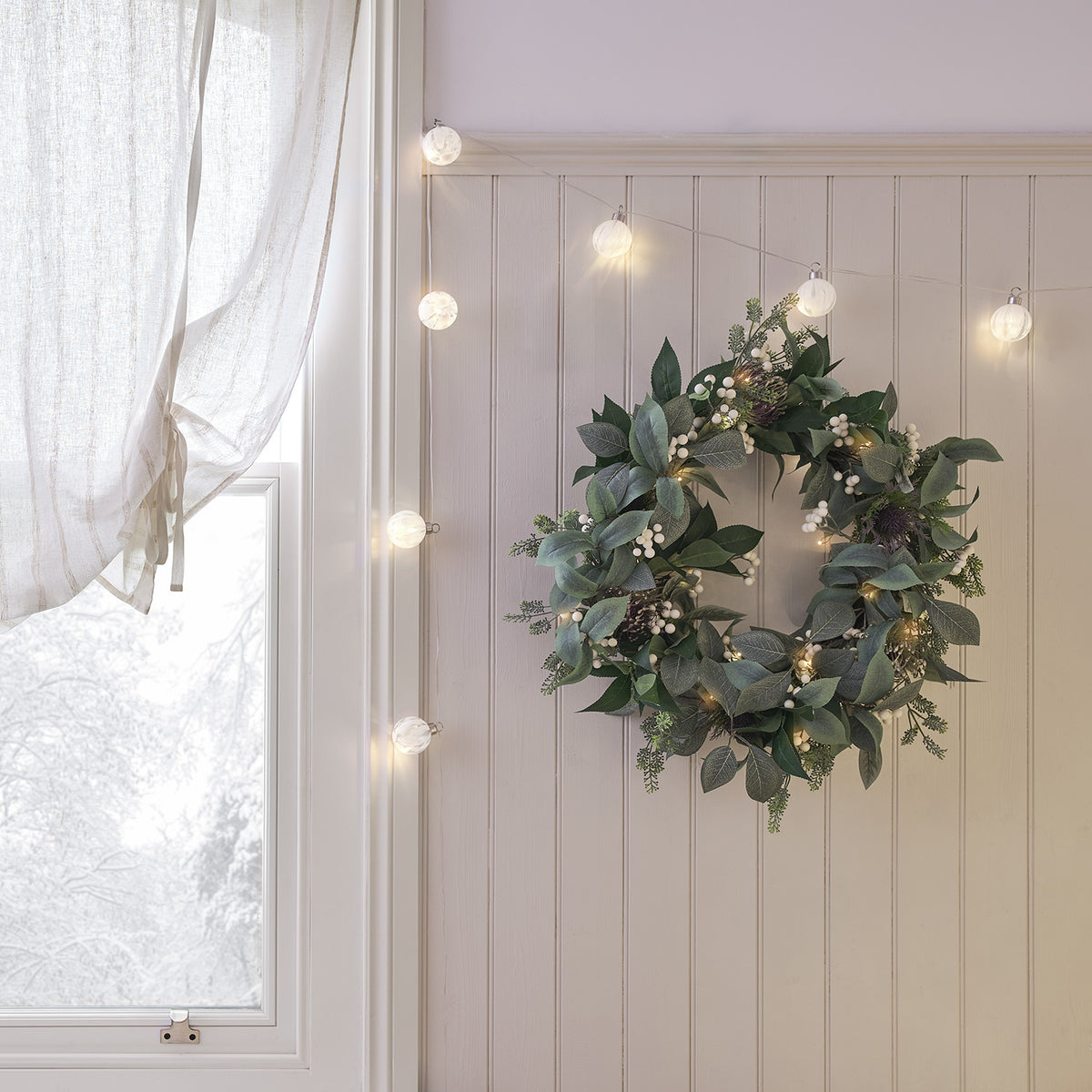 LED Lichterkette mit Weihnachtsbaumkugeln um einen grünen Kranz an weißer Wand drapiert