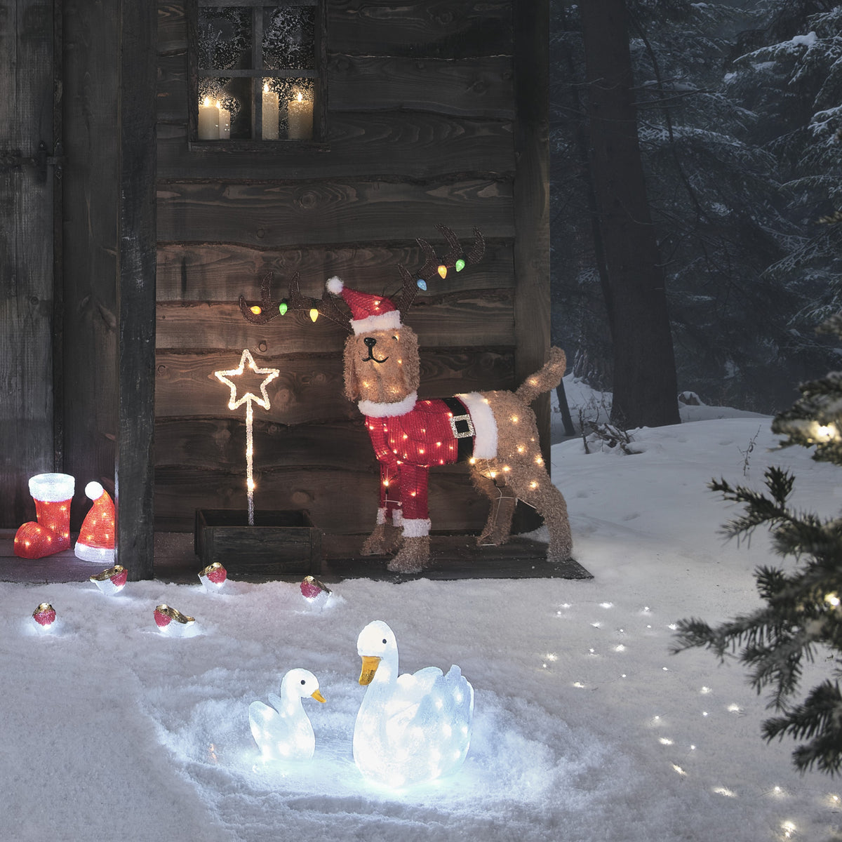 Weihnachtlicher Hund mit Santa-Kostüm und buntem Geweih vor Holzhütte bei Nacht
