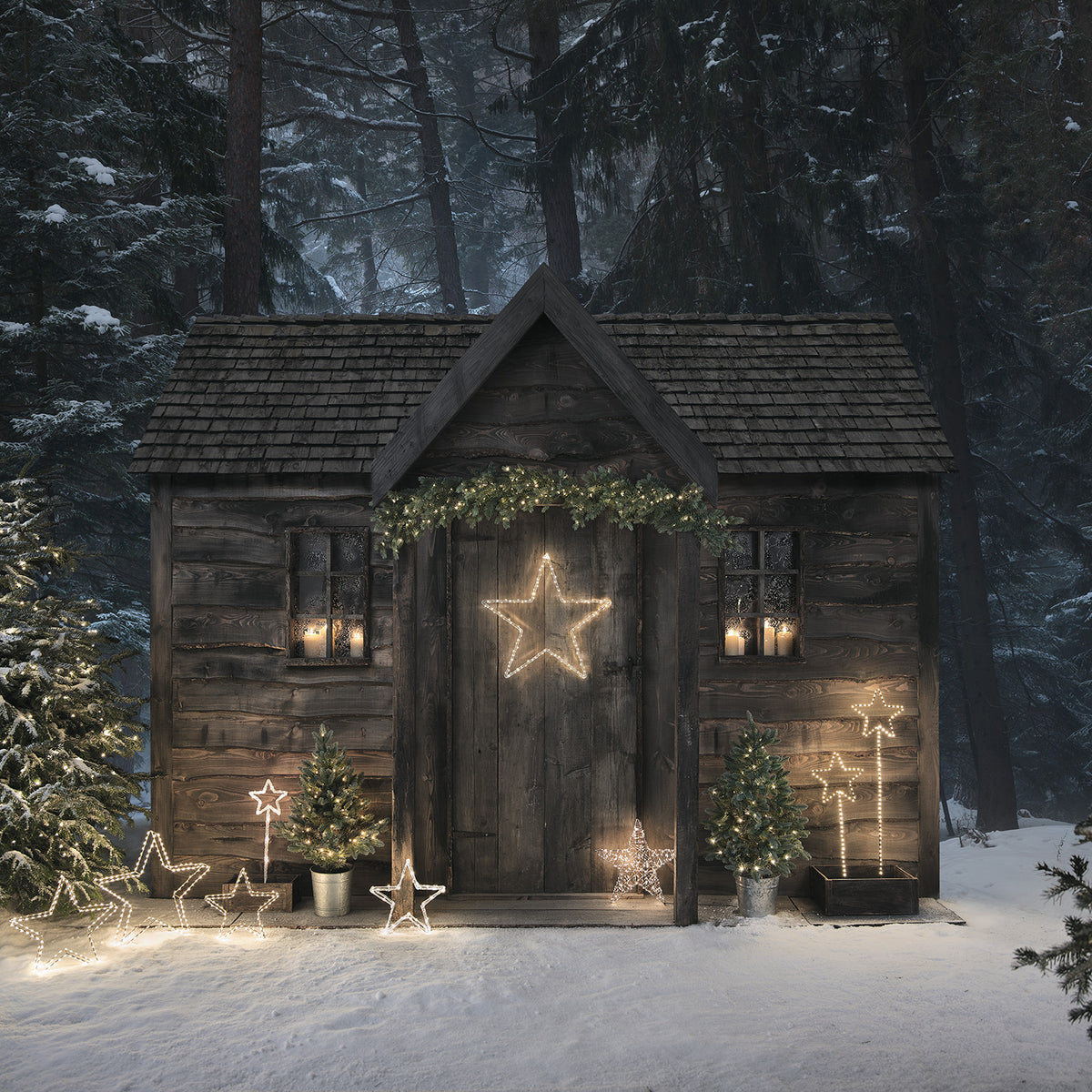 Verschneite Holzhütte mit mehreren leuchtenden Micro LED Sternen und Tannenbäumen vor dem Eingang