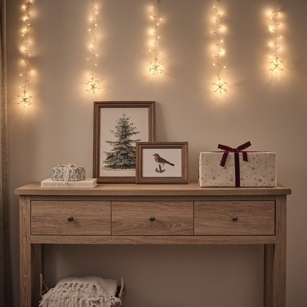 Goldener LED Lichtervorhang mit Starburst-Clustern über Holzkommode mit Weihnachtsbildern und Geschenk