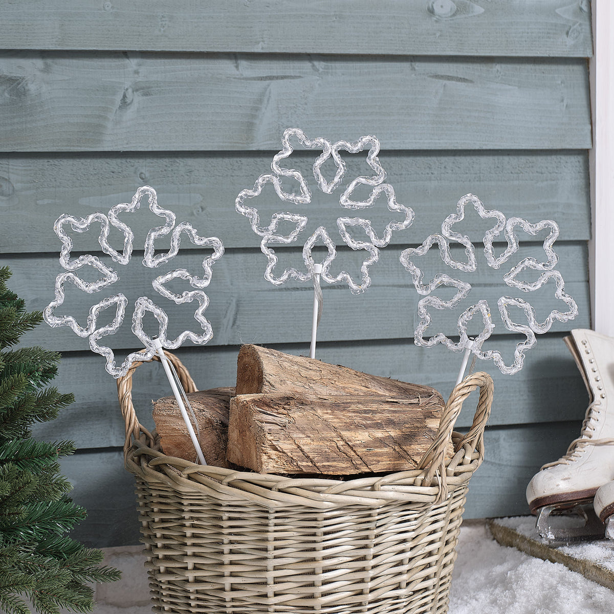 Drei unbeleuchtete Acryl-Schneeflocken Gartenstecker in einem Korb mit Holzscheiten vor Holzwand