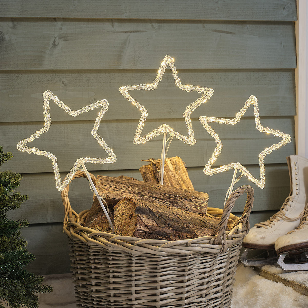 Drei leuchtende Acryl LED Sterne auf Metallstäben in einem Korb mit Holzscheiten vor einer Holzwand