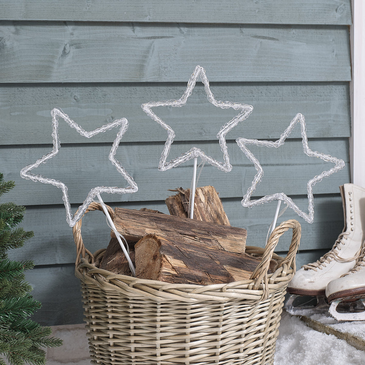 Drei ausgeschaltete Acryl LED Sterne auf Metallstäben in einem Korb mit Holzscheiten vor einer Holzwand