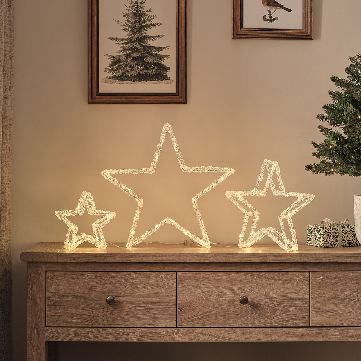 Drei leuchtende LED-Acrylsterne in verschiedenen Größen auf einem Holztisch vor Wand mit Bildern
