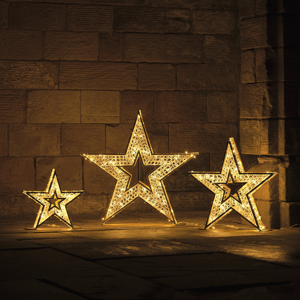 Drei beleuchtete goldene Sterne mit warmweißen LEDs vor dunkler Steinwand bei Nacht