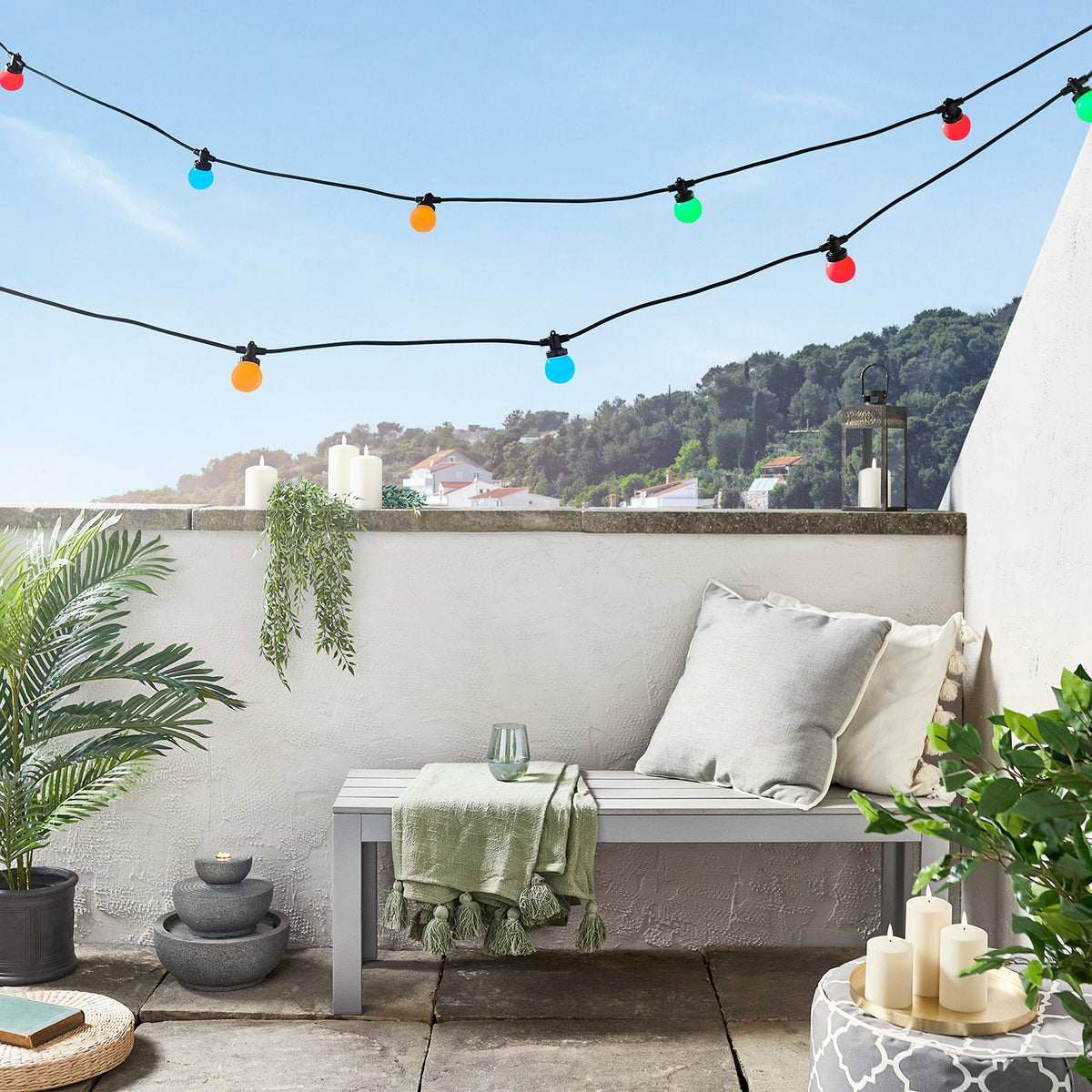 Bunte Lichterkette an schwarzem Kabel auf Terrasse mit Bank, Kissen und Kerzen
