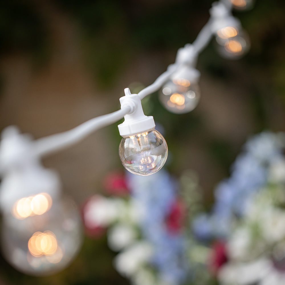 Nahaufnahme einer warmweißen LED-Glühbirne an weißem Kabel vor unscharfem Gartenhintergrund