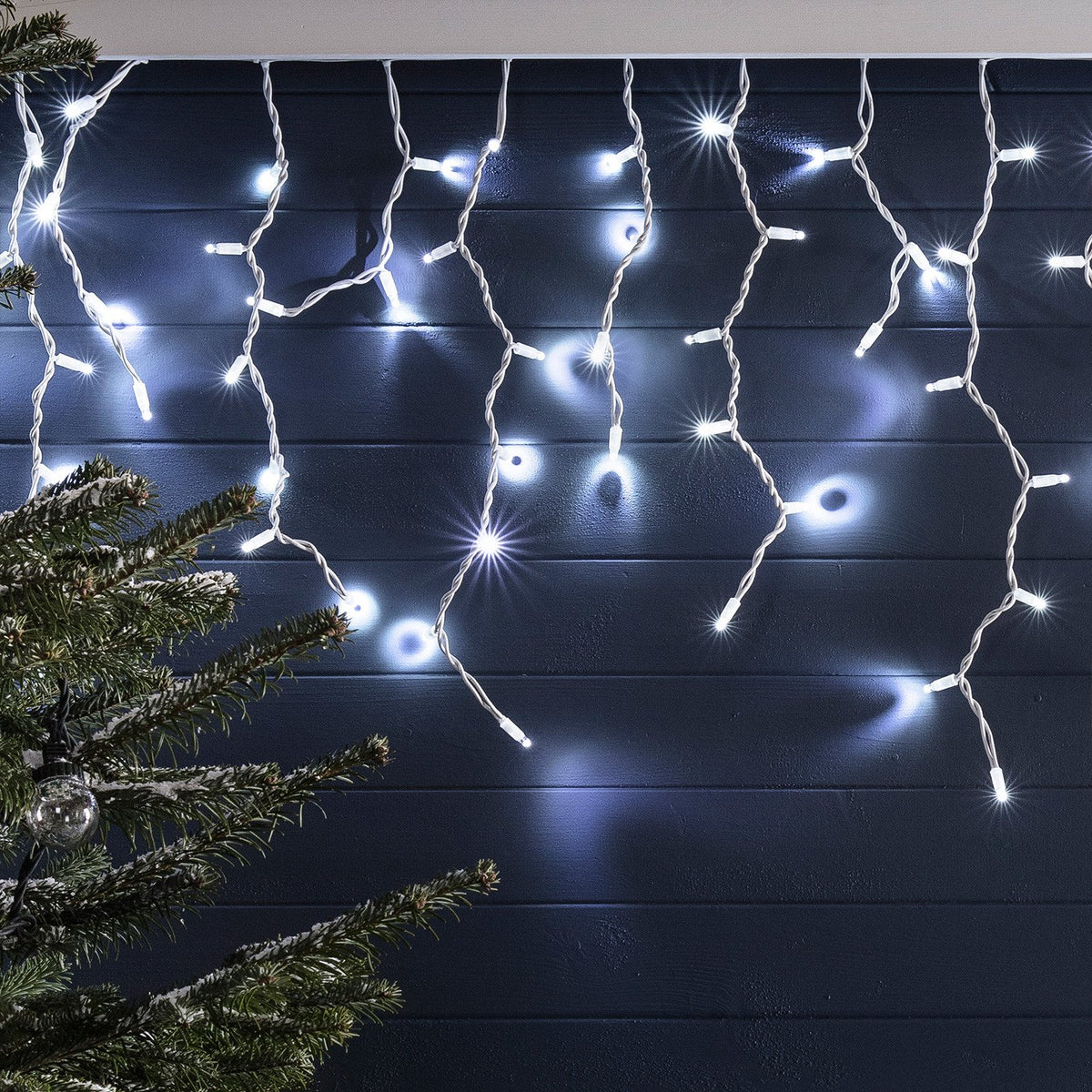 Weiße LED Eiszapfen Lichterkette an dunkler Wand mit Tannenzweig im Vordergrund