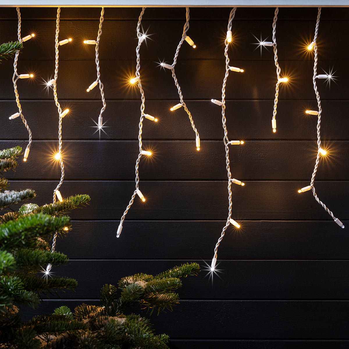 Leuchtende LED Eiszapfen-Lichterkette vor dunkler Holzfassade mit Tannenzweigen