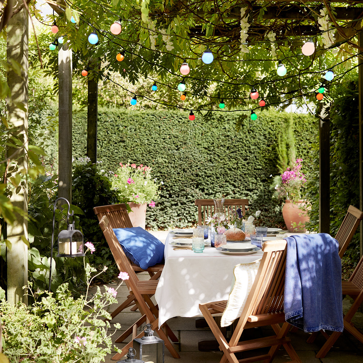 Bunte Lichterkette hängt über gedecktem Gartentisch unter Pergola mit Pflanzen