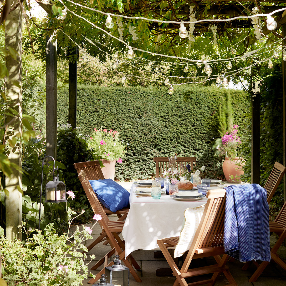 Lichterkette mit warmweißen Glühbirnen über gedecktem Gartentisch unter Pergola