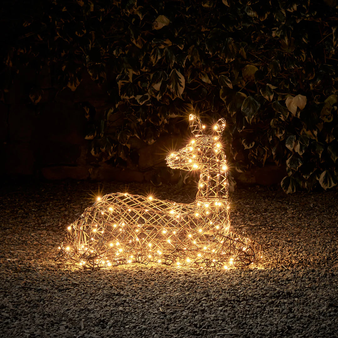 Liegendes LED-Rentier in brauner Rattanoptik mit warmweißen Lichtern bei Nacht im Garten