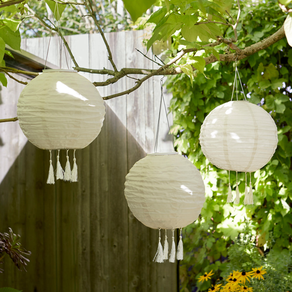 Drei cremefarbene Solar-Lampion mit Fransen hängen an Ästen im Garten