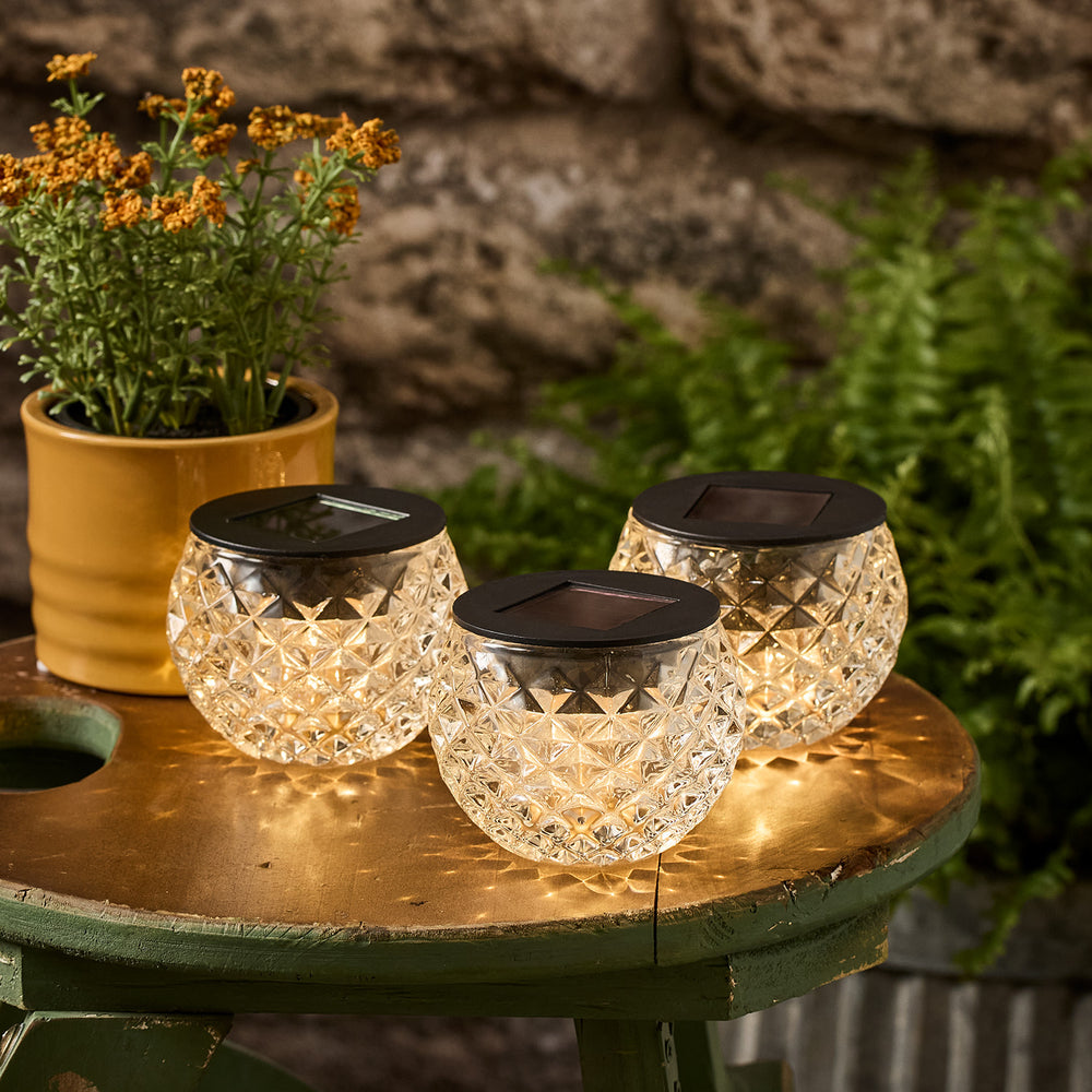 Drei solarbetriebene Windlichter aus strukturiertem Glas auf einem Holztisch im Garten