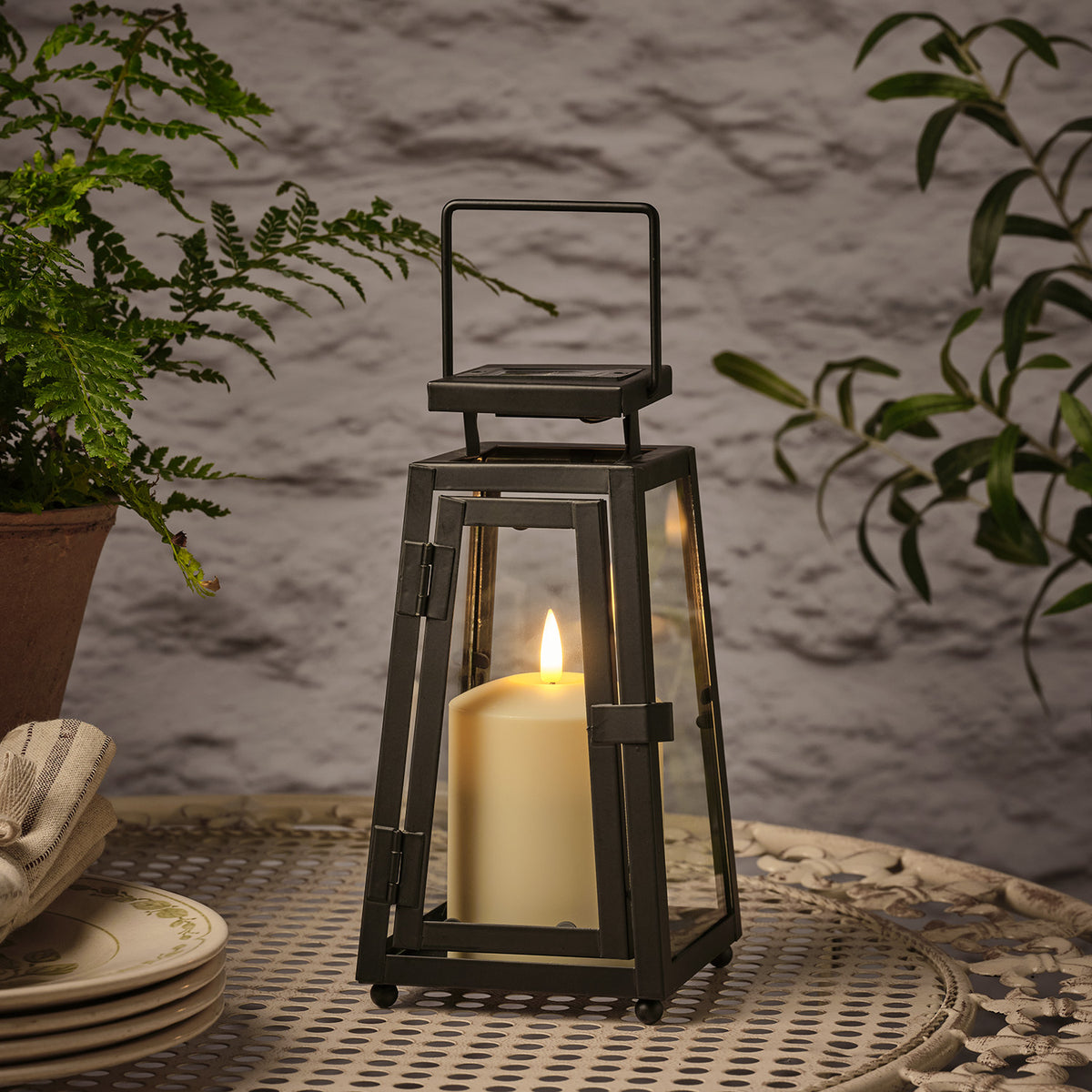 Schwarze Solarlaterne mit flackernder TruGlow® LED-Kerze auf Metalltisch neben Pflanzen