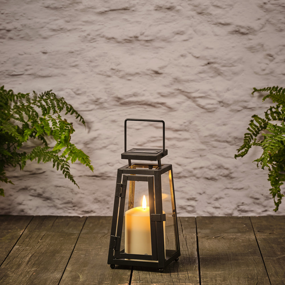 Schwarze Solarlaterne mit TruGlow® LED-Kerze auf Holzterrasse zwischen grünen Pflanzen