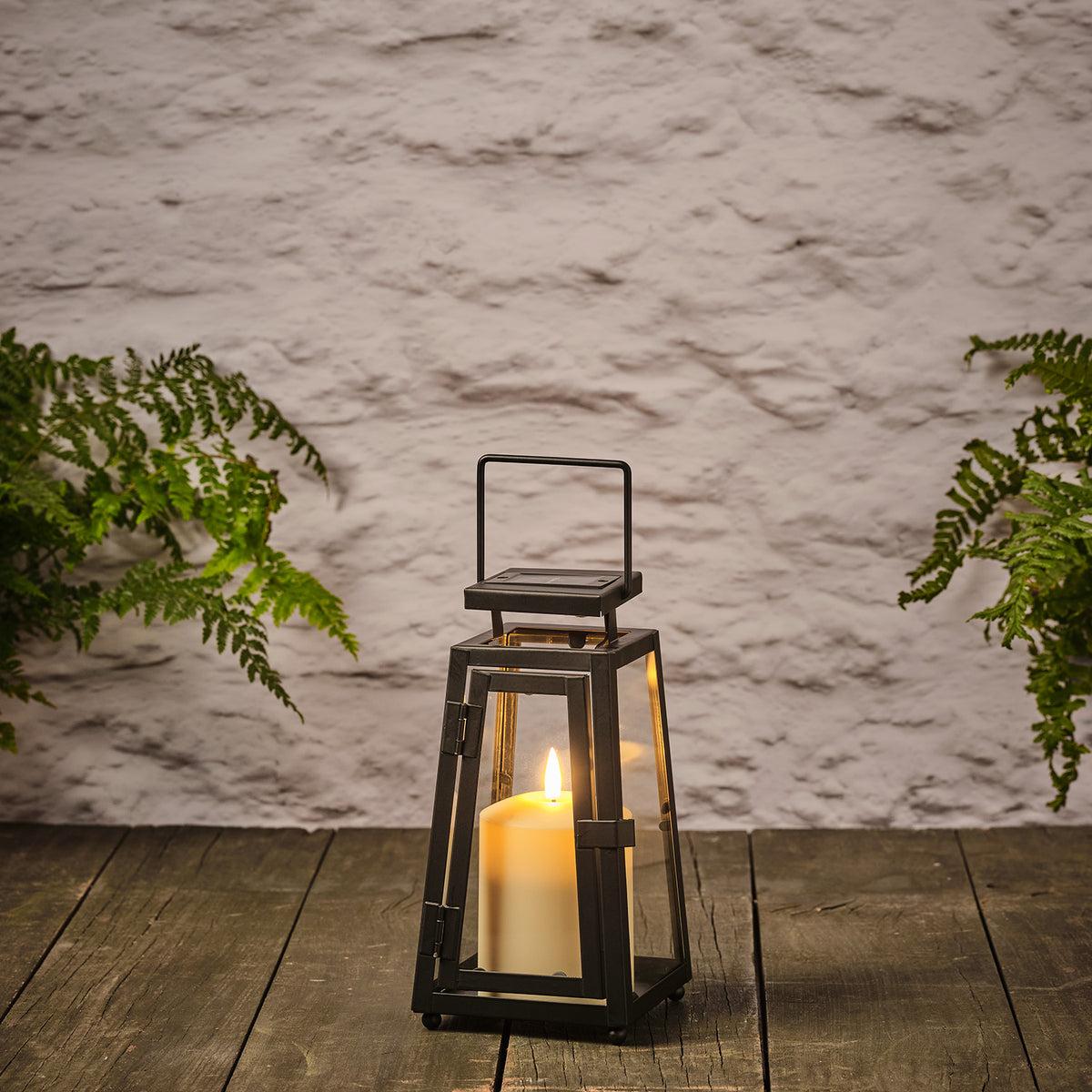Schwarze Solarlaterne mit TruGlow® LED-Kerze auf Holzterrasse zwischen grünen Pflanzen