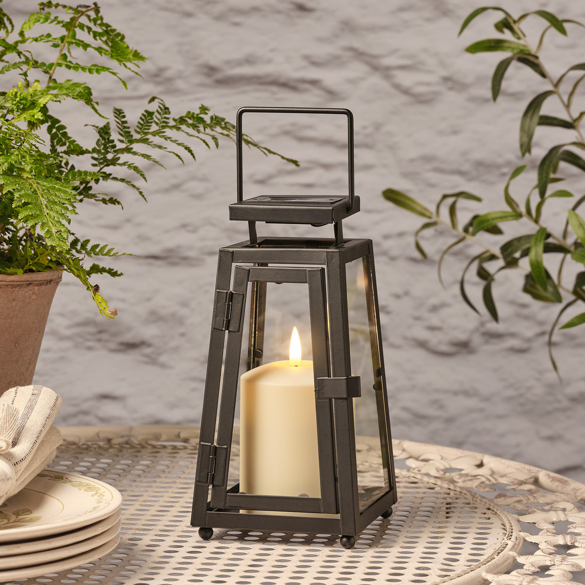 Schwarze Solarlaterne mit flackernder TruGlow® LED-Kerze auf Metalltisch neben Pflanzen