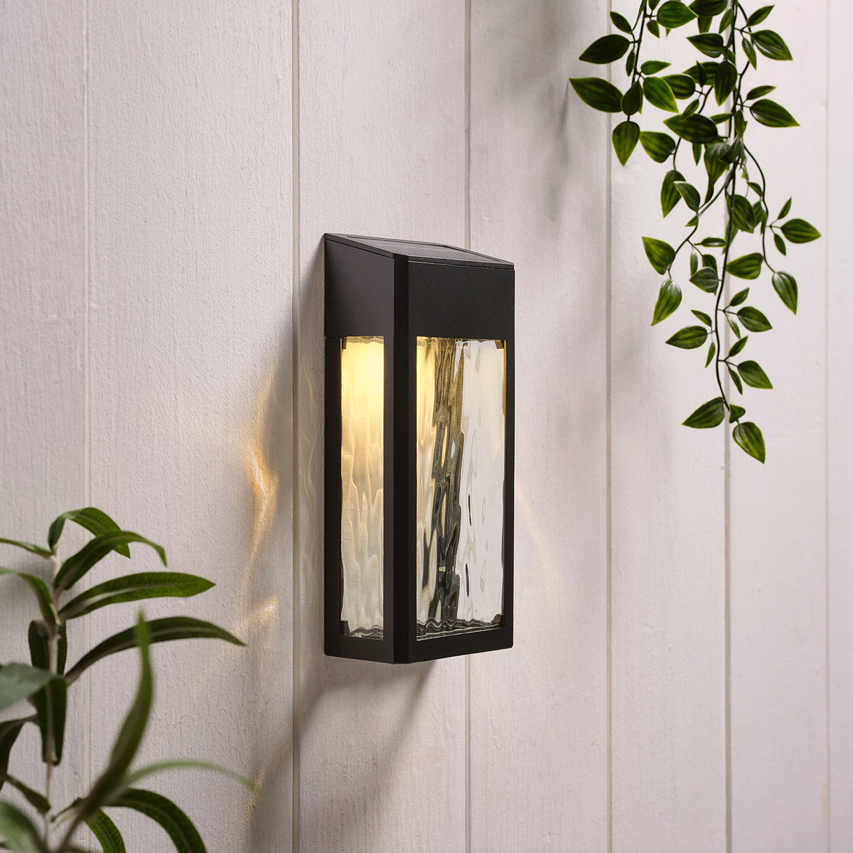 Solar-Wandleuchte mit warmweißem Licht an weißer Holzpaneelwand neben grünen Blättern