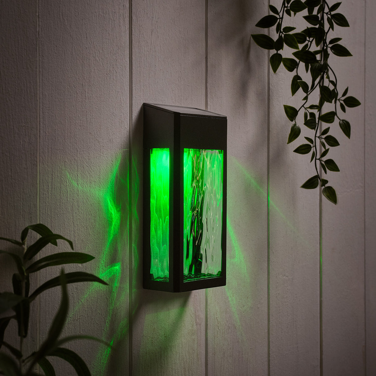Solar-Wandleuchte mit grünem Licht an weißer Holzpaneelwand mit Pflanzenblättern