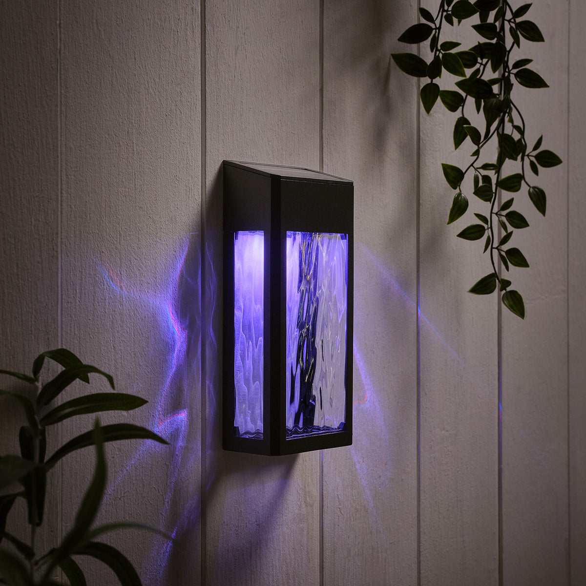 Schwarze LED Solar Wandleuchte mit strukturiertem Glas leuchtet in blauem Farbwechsel an weißer Wand