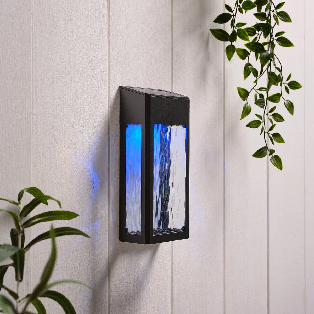Solar-Wandleuchte mit blauem Licht an weißer Holzpaneelwand mit Pflanzenblättern
