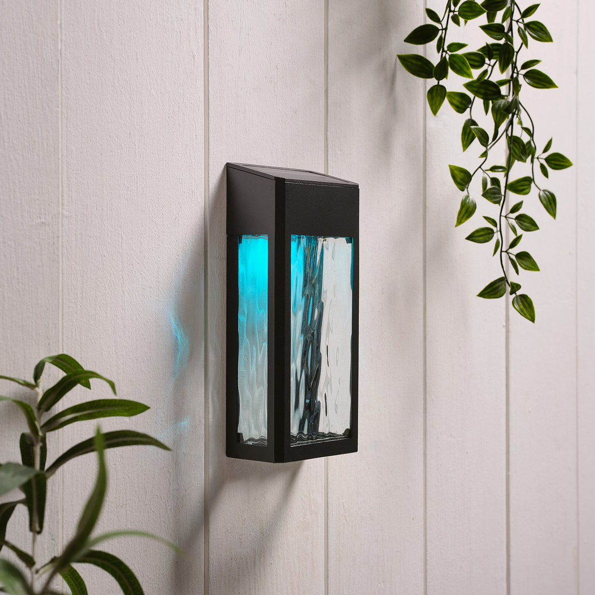 Solar-Wandleuchte mit hellblauem Licht an weißer Holzpaneelwand neben grünen Blättern