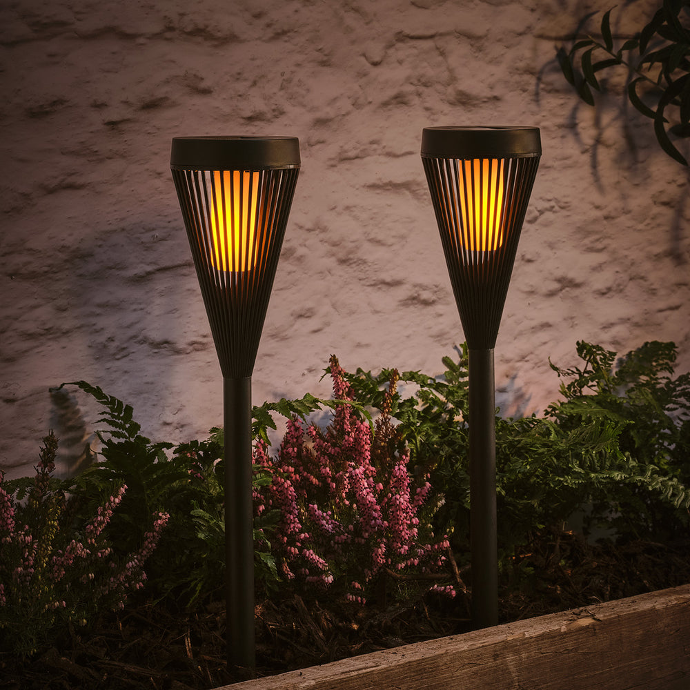 Zwei schwarze Solar-Gartenfackeln mit flackernder LED-Flamme im Beet vor einer Wand