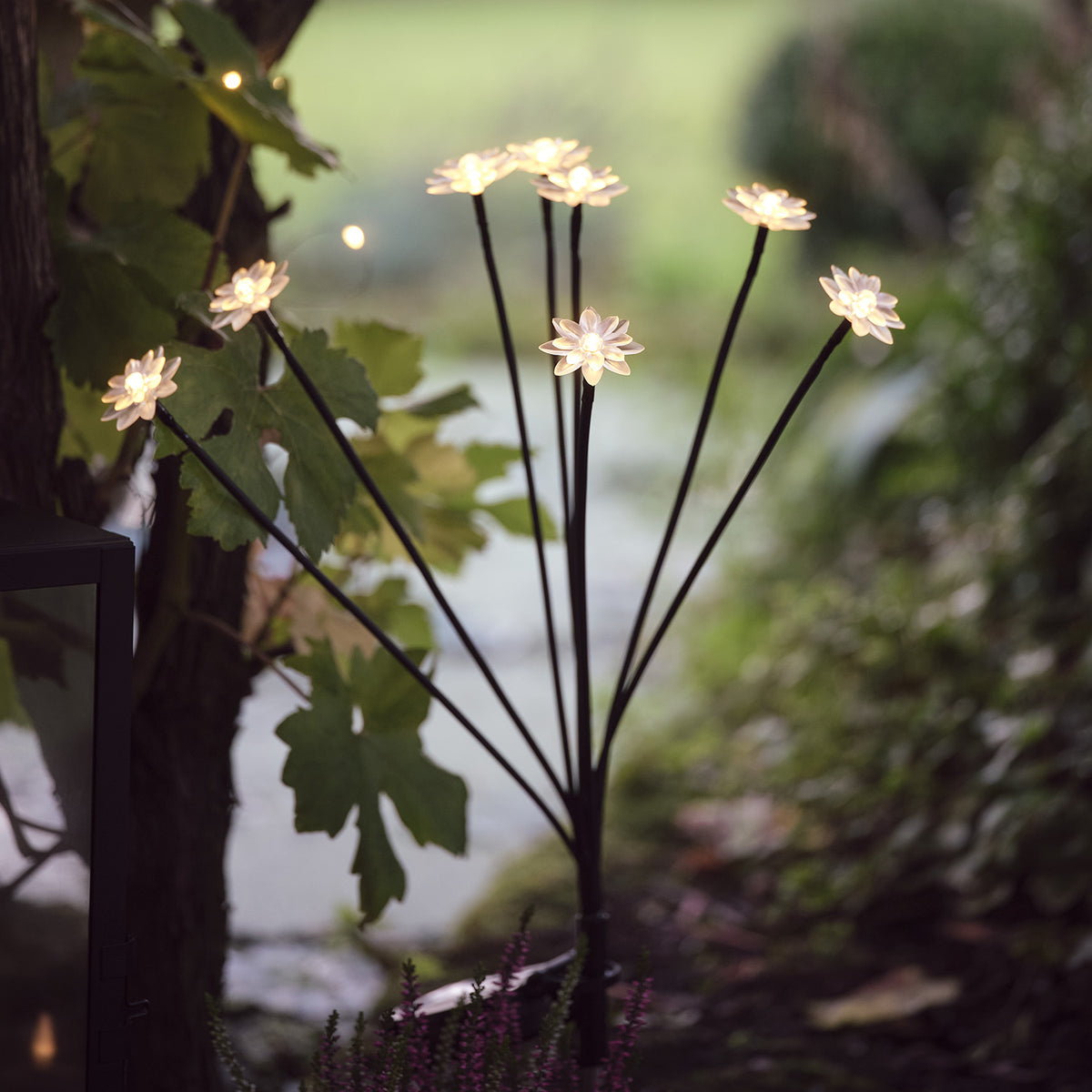 Acht Solar-Blumenleuchten auf schwarzen Stangen im Garten bei Dämmerung mit grünen Blättern