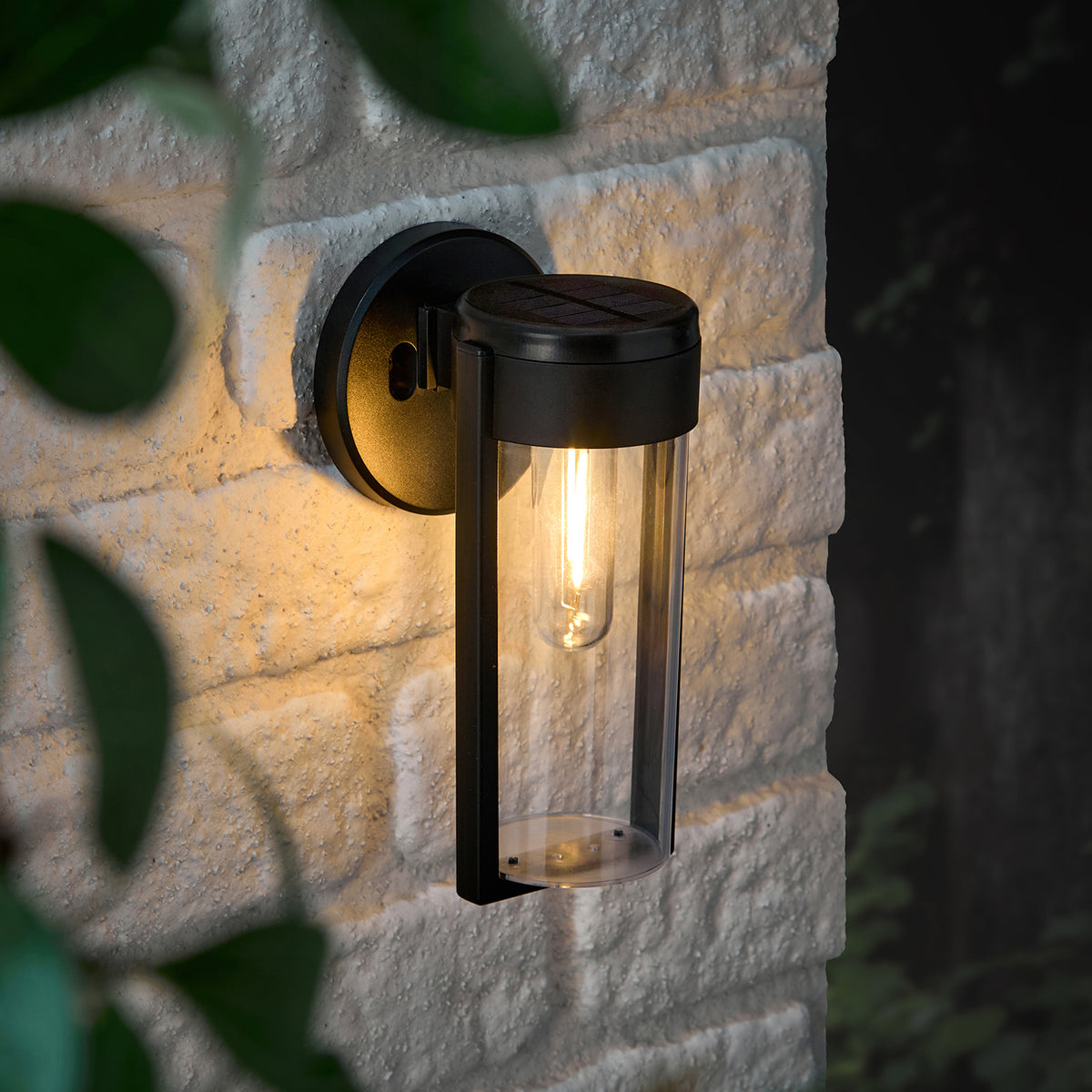 Schwarze Solar-Wandleuchte Wilton mit warmweißem Licht an heller Steinwand bei Dämmerung