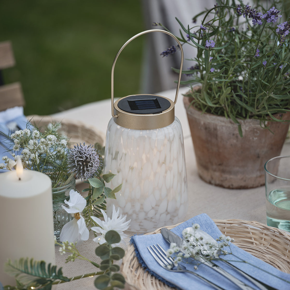 Solarlaterne mit goldfarbenem Henkel und Deckel auf Tisch mit Blumen und blauem Stoffserviette