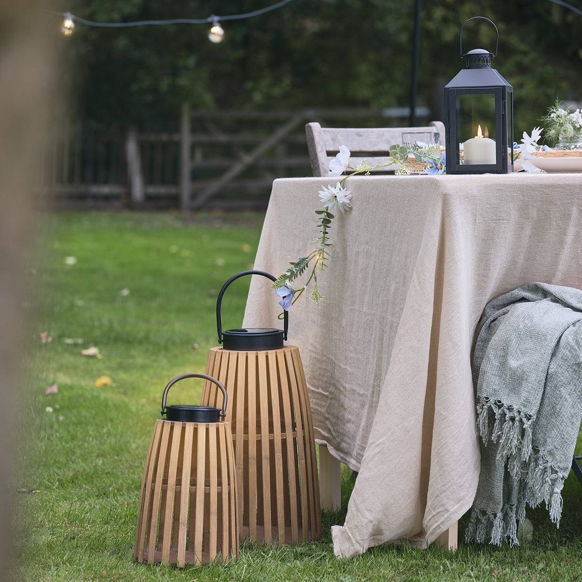 Zwei Bambus Solarlaternen neben Gartentisch mit Leinentischdecke und Decke auf Stuhl