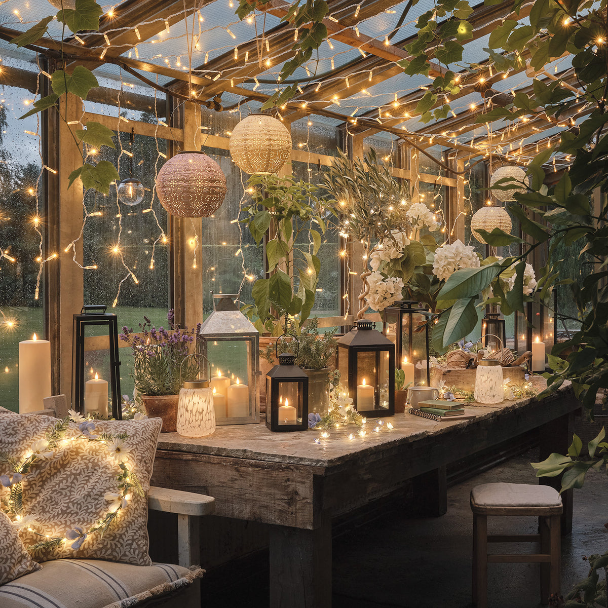 Gemütlich beleuchteter Wintergarten mit verschiedenen Laternen und Lichterketten an Holztisch und Decke
