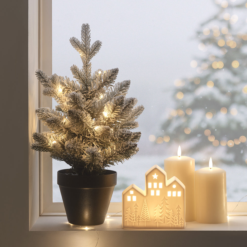 Verschneiter Mini-Tannenbaum mit warmweißen LEDs und beleuchtetes Keramikhäuschen auf Fensterbank