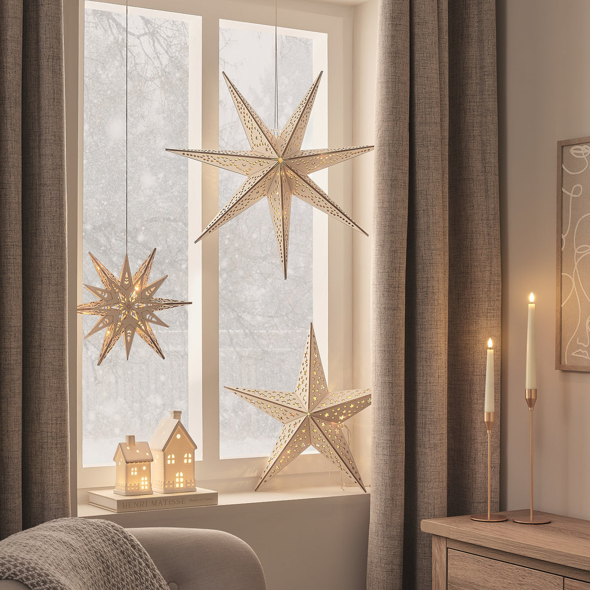 Wohnzimmerfenster mit drei beleuchteten Holzsternen, Kerzen und dekorativen Häuschen