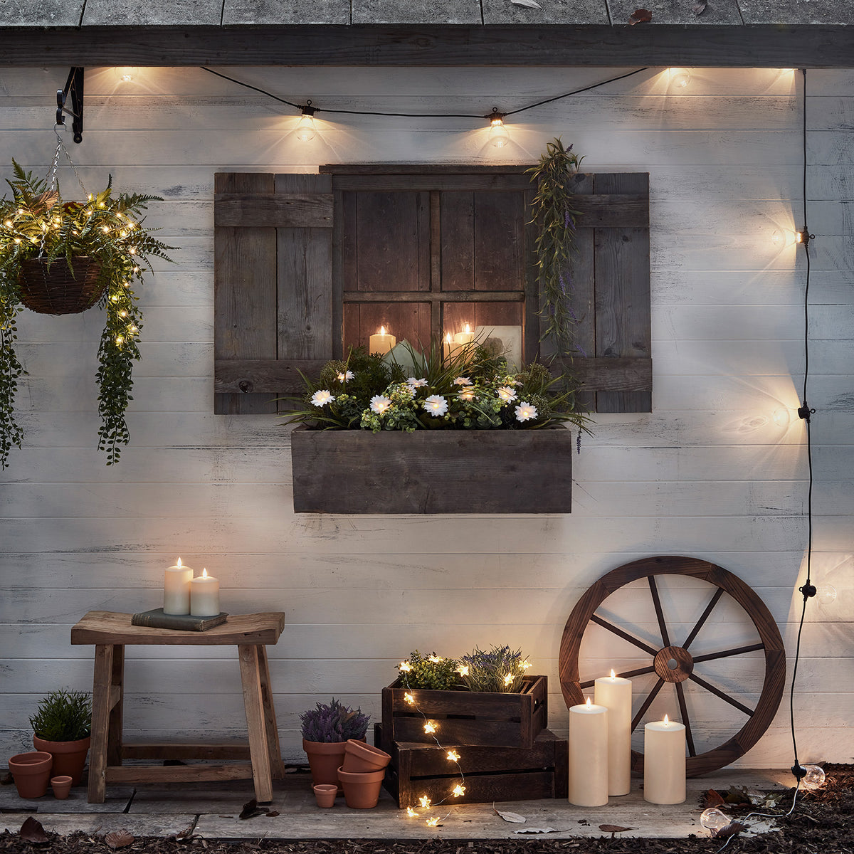 Lichterkette mit warmweißen Glühbirnen dekoriert an weißer Holzfassade mit Pflanzen und Kerzen