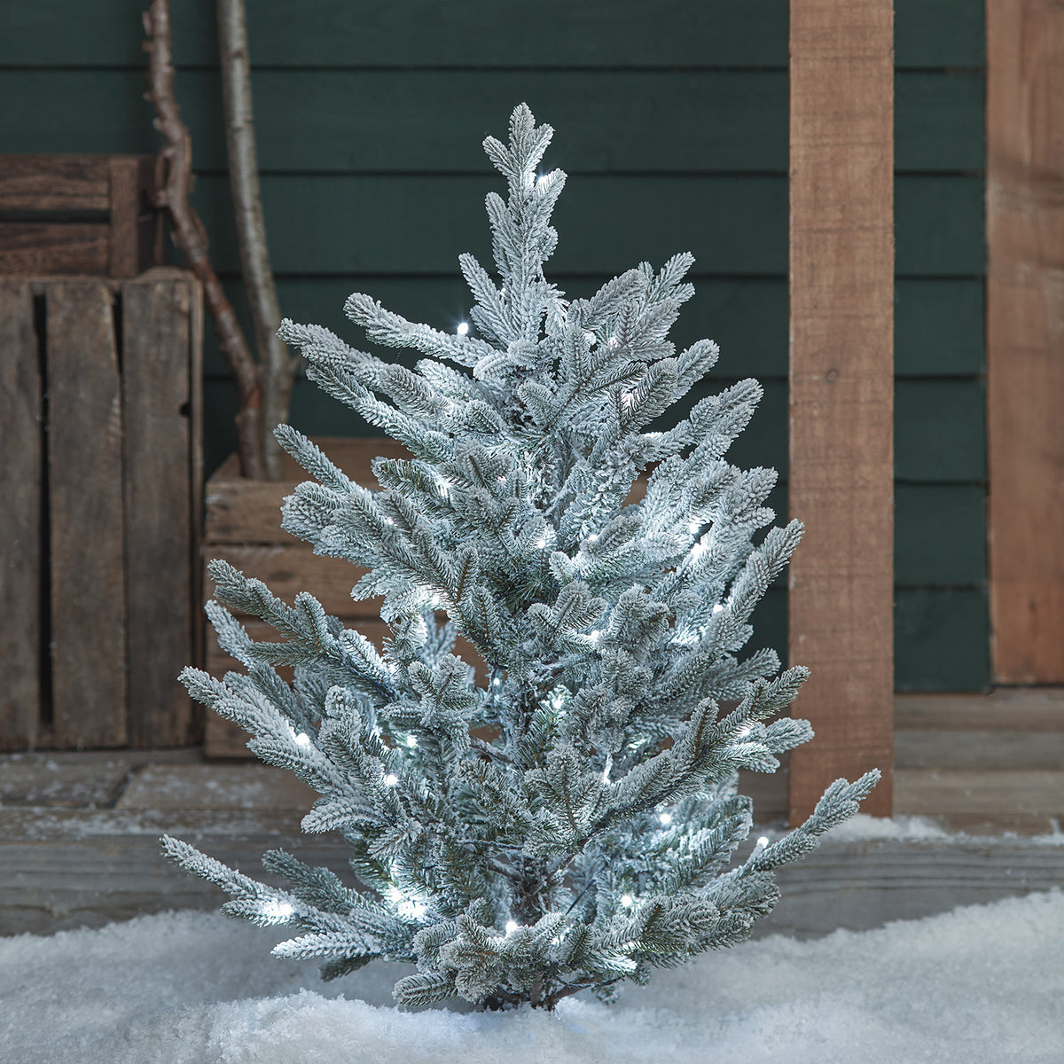 Verschneiter Weihnachtsbaum mit kaltweißen LED-Lichtern im Schnee vor Holzverkleidung