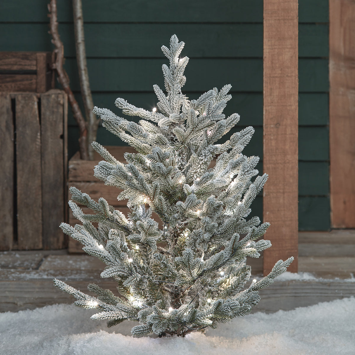 Verschneiter Weihnachtsbaum mit weißen LED-Lichtern im Schnee vor Holzverkleidung