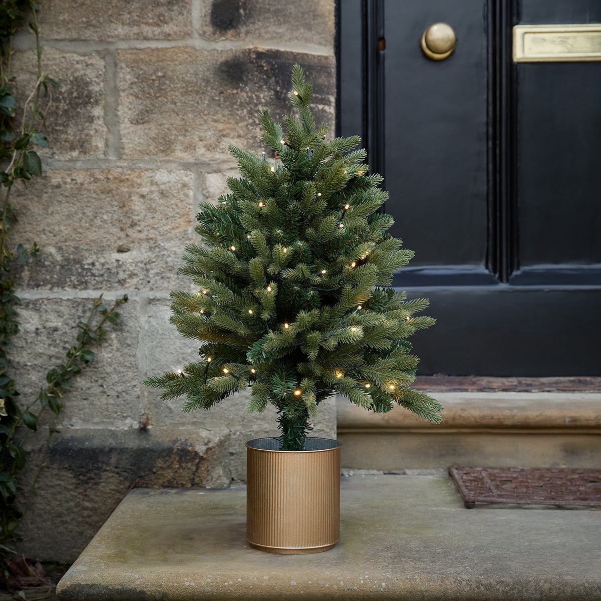 Beleuchteter 75 cm hoher Weihnachtsbaum im goldenen Topf vor einer Steinwand und schwarzer Tür