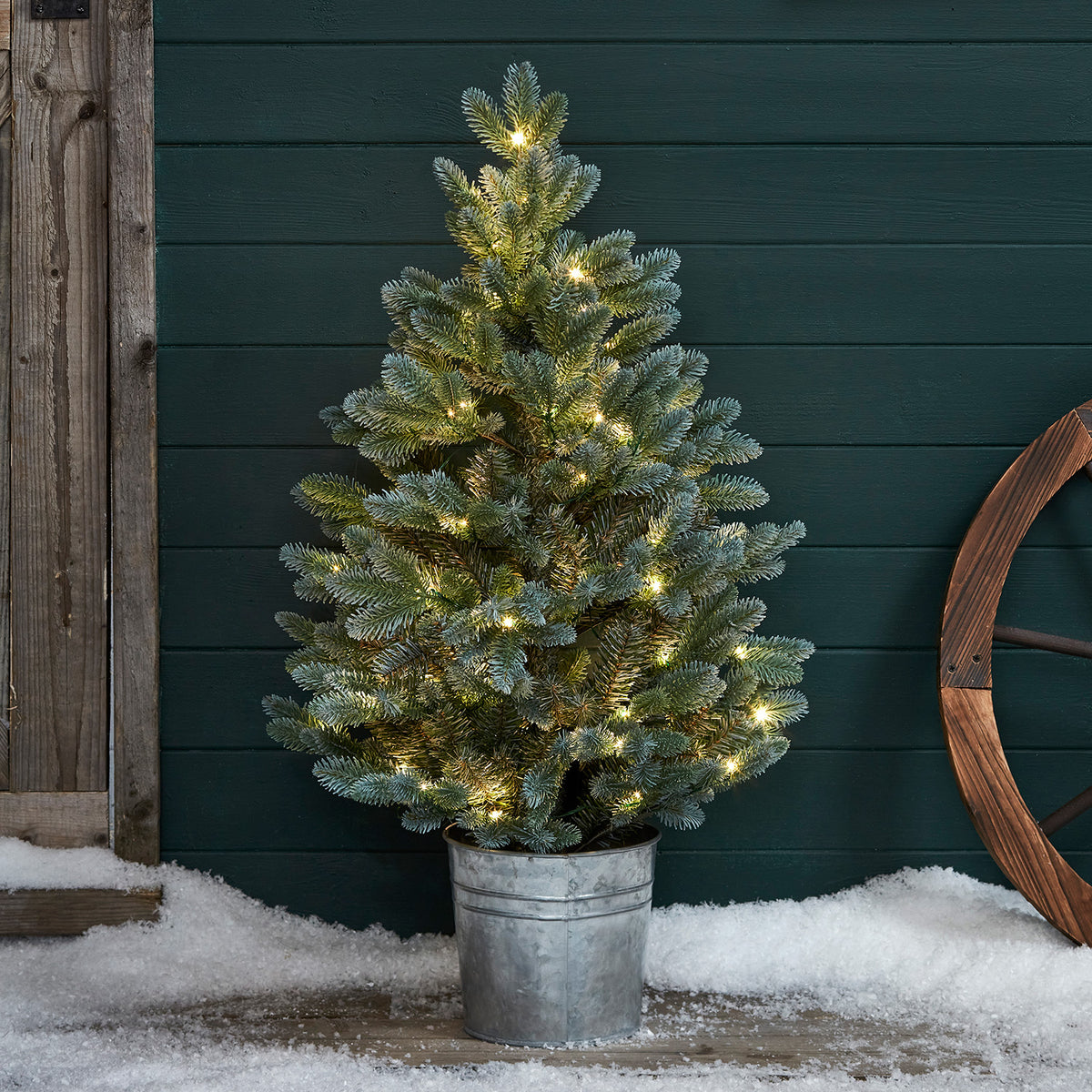 Künstlicher Weihnachtsbaum mit warmweißen LEDs im Metalltopf vor grüner Wand und Schnee
