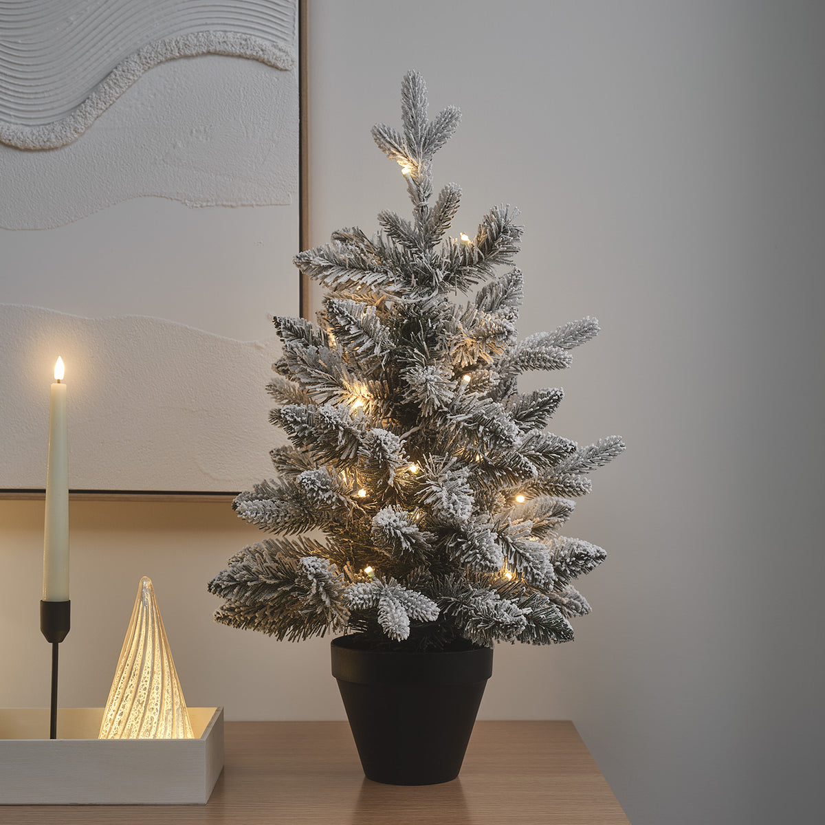 Beleuchteter verschneiter LED-Weihnachtsbaum auf Holztisch neben Kerze und Glasdeko