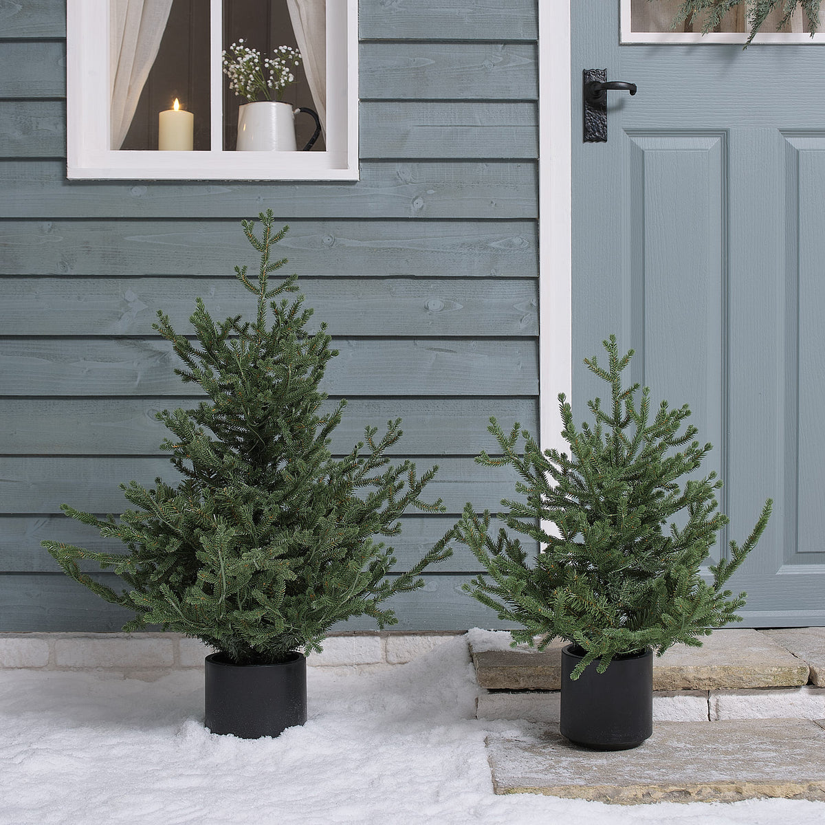 Zwei künstliche Weihnachtsbäume im Topf ohne Beleuchtung auf Schnee vor Hauswand