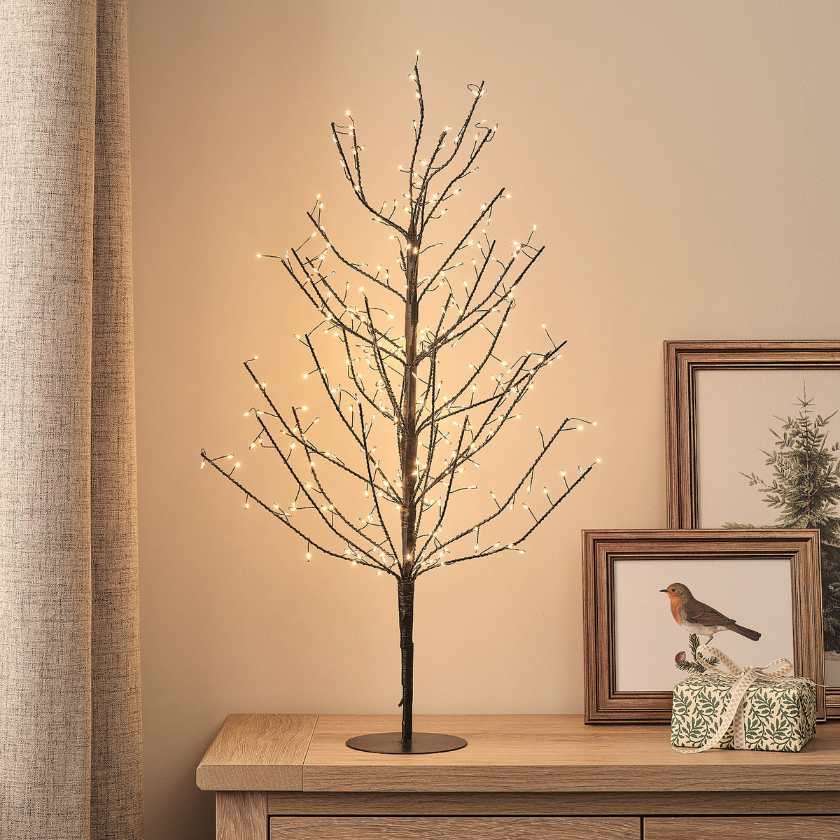 Schwarzer LED-Lichterbaum mit 300 warmweißen Mikro-LEDs auf Holzkommode neben Bilderrahmen und Geschenk