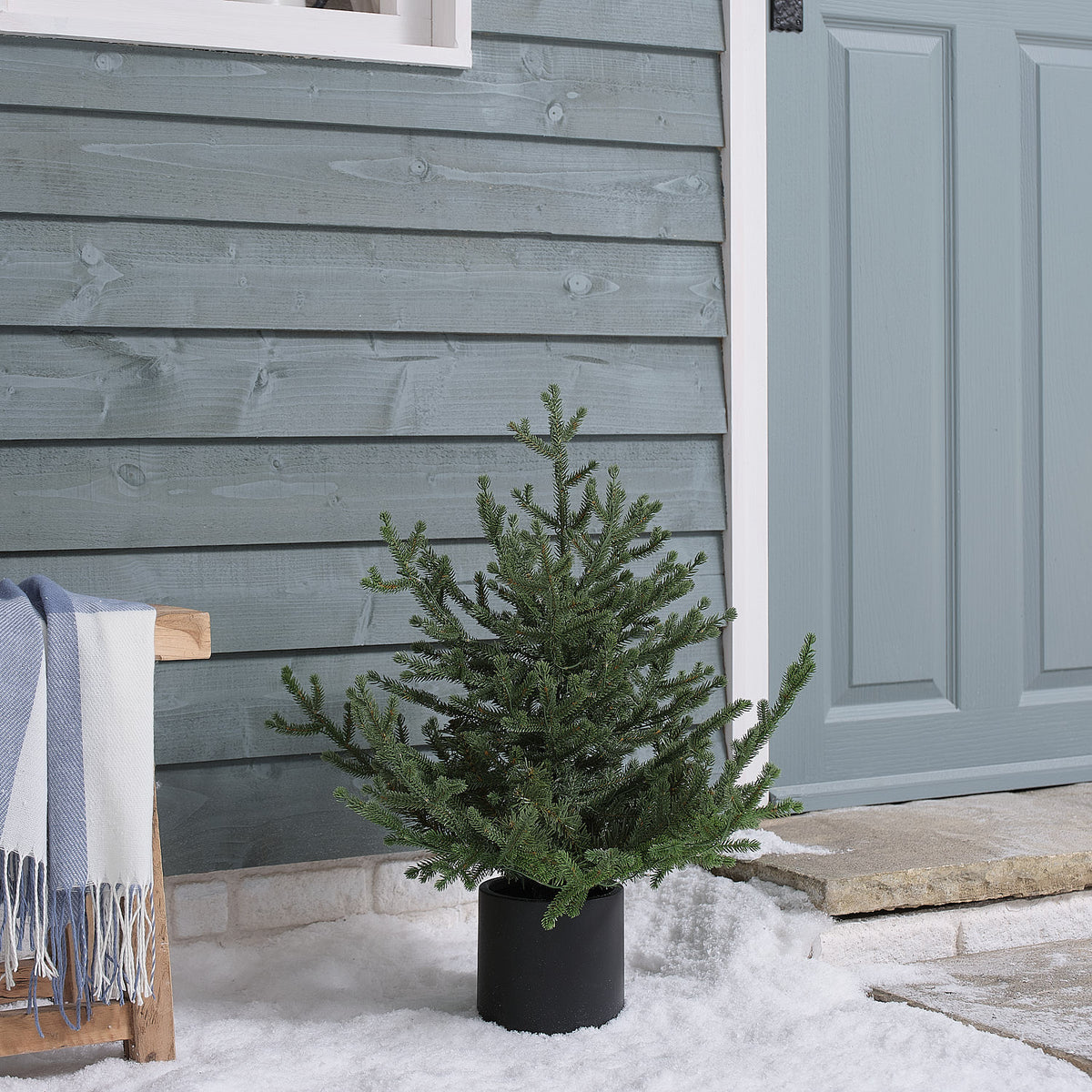 Unbeleuchteter künstlicher Weihnachtsbaum im schwarzen Topf vor Hauswand mit Schnee