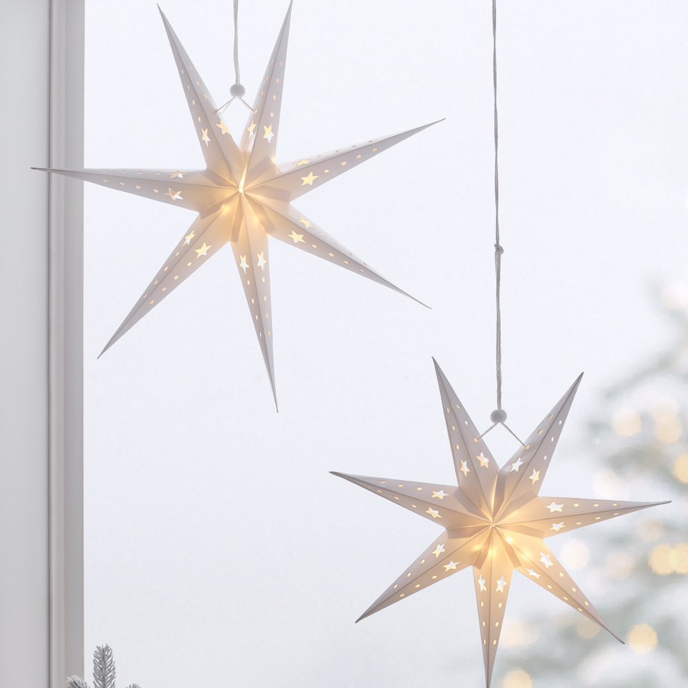 Zwei weiße Papiersterne mit LED-Beleuchtung hängen vor einem Fenster mit winterlicher Außenansicht