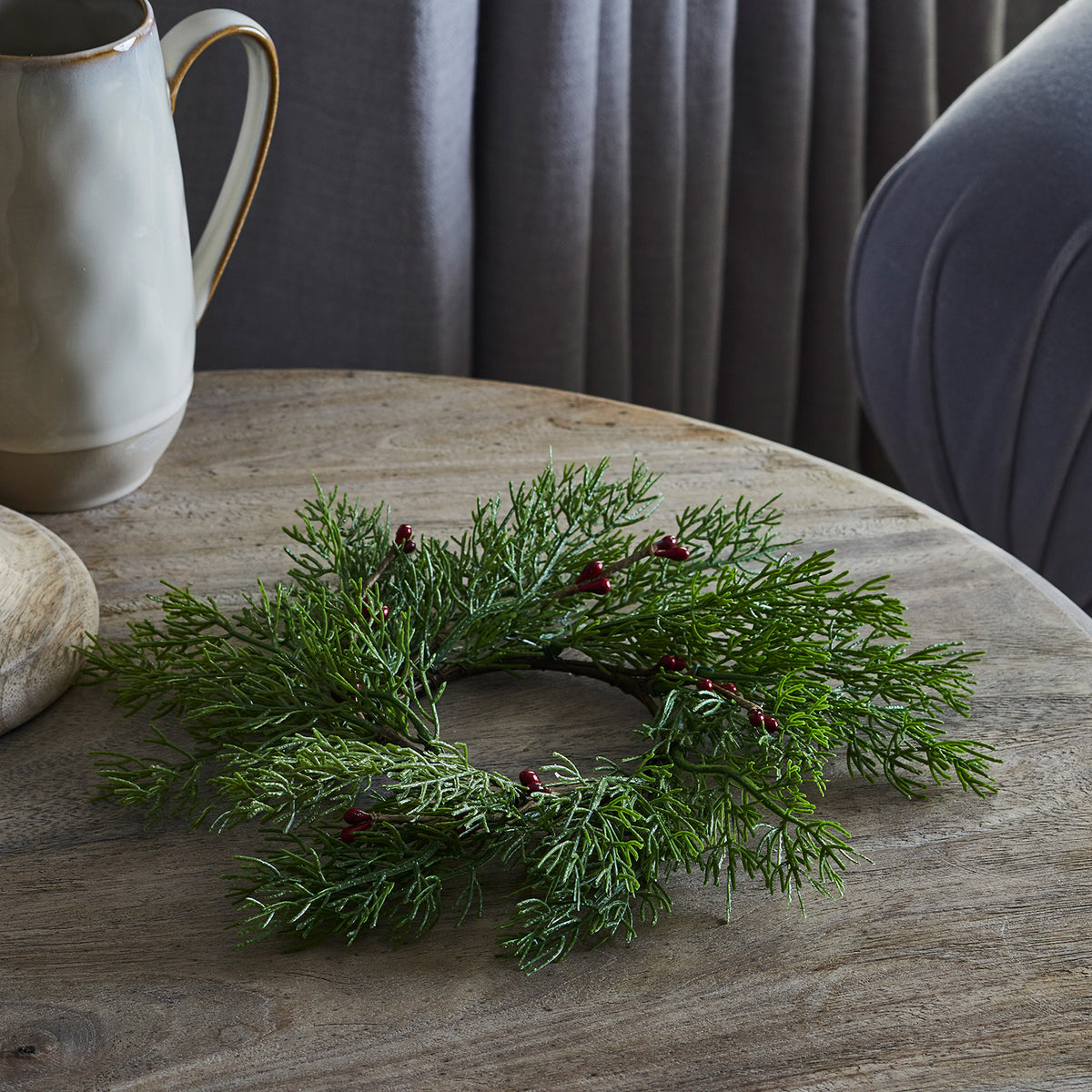 Weihnachtskranz mit grünen Zweigen und roten Beeren auf Holztisch neben grauem Sessel
